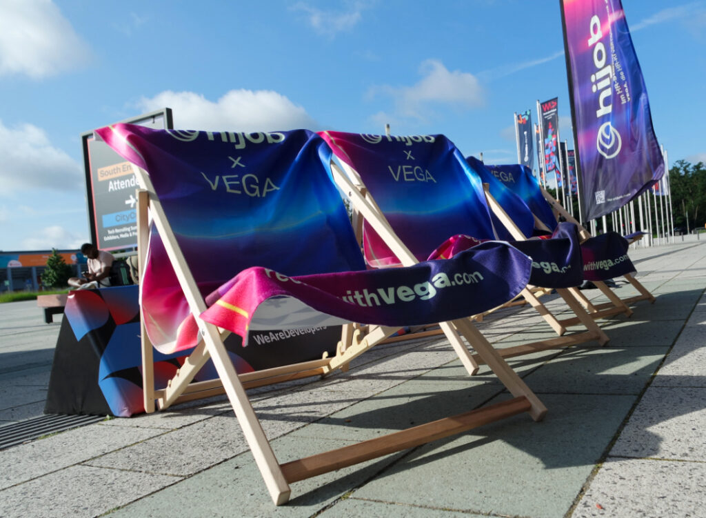 Es sind Liegestühle mit lila-blauen Handtüchern von hijob x Vega zu sehen. Im Hintergrund weht ein Banner von hijob und sowie weitere Banner der Messe.